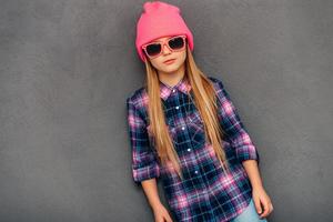 Confident little miss. Beautifullittle girl in sunglasses and beanie hat looking at camera while standing against grey background photo