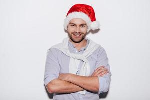 Merry Christmas Handsome young man in Santa hat keeping arms crossed and smiling while standing against white background photo