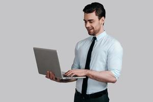 Using modern technologies. Handsome young man in formalwear holding laptop and typing something while standing against grey background photo