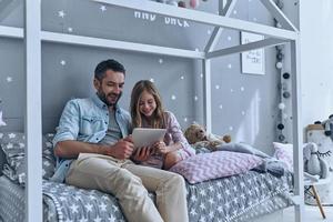 Using technologies for fun. Young father and his little daughter looking at digital tablet while sitting on the bed at home photo