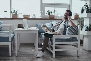 Good talk. Handsome young man in eyewear talking on smart phone and smiling while sitting on the sofa photo