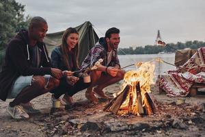 Totally happy. Group of young people in casual wear roasting marshmallows over a campfire while resting near the lake photo