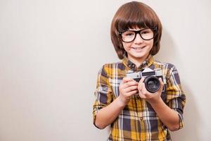 joven fotógrafo. niño pequeño con anteojos sosteniendo la cámara y sonriendo mientras está de pie contra el fondo gris foto