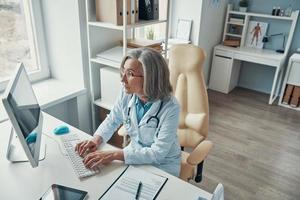 Top view of mature female doctor in white lab coat working on computer while sitting in her office photo