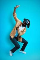 Full length of playful young African man wearing virtual reality glasses while standing against blue background photo