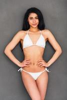 Looking gorgeous. Beautiful young mixed race woman in white bikini holding hands on hip and smiling while standing against grey background photo