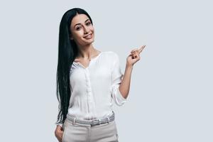 Thinking about new solutions. Young beautiful woman in white shirt pointing away and smiling while standing against grey background photo