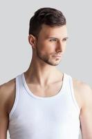 Confident handsome. Handsome young muscular man in white tank top looking away while standing isolated on grey background photo