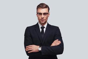 Portrait of confidence. Handsome young man in full suit keeping arms crossed and looking at camera while standing against grey background photo