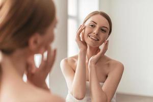 Beauty and her reflection in a mirror. Over the shoulder view of attractive woman touching her face and smiling while spending time at home photo