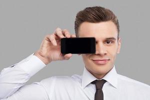 Man with mobile. Handsome young man in shirt and tie holding mobile phone in front of the eye and smiling while standing against grey background photo