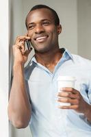 Relaxing during coffee break. Cheerful young African man in blue shirt holding a cup of coffee and talking on the mobile phone photo