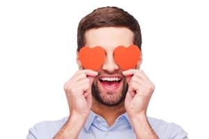 This is love Handsome young man holding heart shaped valentine cards in front of his eyes and smiling while standing isolated on white background photo