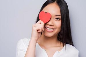 Look around, and you will find you love Beautiful young Asian woman holding heart shaped valentine card in front of her eye and smiling while standing against grey background photo