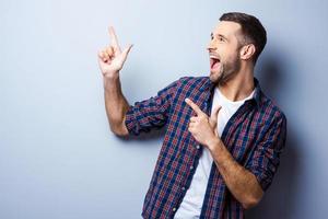 Just look at that Happy young man in casual shirt pointing away and smiling while standing against grey background photo
