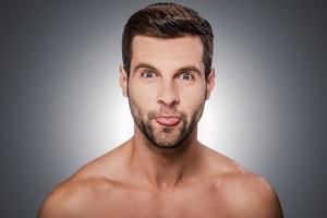 mueca graciosa. retrato de un apuesto joven sin camisa mirando a la cámara y mostrando su lengua mientras se enfrenta a un fondo gris foto