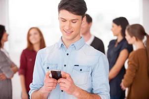 compartir buenas noticias con amigos. joven alegre sosteniendo un teléfono móvil y sonriendo mientras un grupo de personas se comunica en segundo plano foto