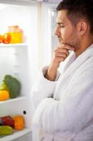 What to eat Thoughtful young man in bathrobe standing near the open fridge and looking inside photo