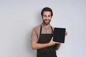 Cheerful young man in apron showing his digital tablet and smiling while standing against gray background photo