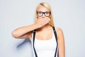 Stop gossip Surprised young woman covering mouth with hand and staring at camera while standing against grey background photo