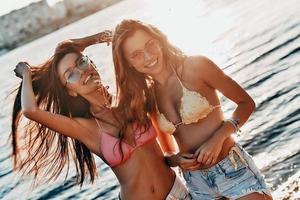 diosas del verano. dos atractivas mujeres jóvenes en traje de baño sonriendo y mirando hacia otro lado mientras están de pie cerca del río al aire libre foto