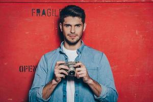 Can I take a picture of you Handsome young man holding retro styled camera and looking camera while standing against red background photo