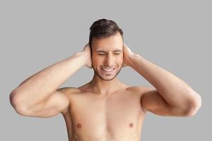 Cant hear it anymore. Handsome young muscular man covering his ears with hands and keeping eyes closed while standing isolated on grey background photo