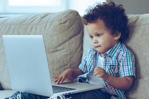 It is time to learn something new.Little African baby boy using his laptop while sitting on the couch at home photo