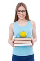 Confident and smart. Cheerful teenage girl holding book stack with apple on it and smiling while standing isolated on white photo