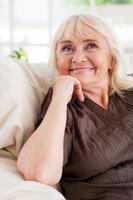 Day dreaming. Thoughtful senior woman holding hand on chin and smiling while sitting in a chair photo