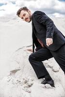 Moving up by desert dune. Confident young man in formalwear rising up by desert dune and looking at camera photo