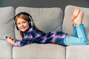 Best free time ever Beautiful little girl in headphones holding her smart phone and looking at camera with smile while lying on front on the couch at home photo