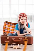 Bored of playing. Bored little boy in pilot headwear and eyeglasses holding hand on chin and looking at camera while sitting inside of briefcase at home photo