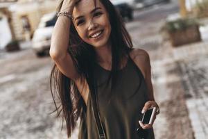 se ve increíble. Atractiva mujer joven manteniendo la mano en el cabello y sonriendo mientras está de pie al aire libre foto