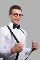 Confident nerd. Cheerful young man in bow tie adjusting his suspenders and smiling at camera while standing against grey background photo