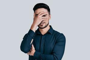 Feeling curious.  Handsome young man covering face with hand and looking at camera while standing against grey background photo