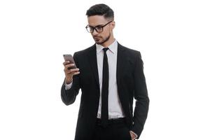 Quick chat with business partner. Serious young man in full suit typing text message using his smart phone while standing against white background photo