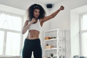 Strong young African woman in sports clothing kickboxing while exercising at home photo