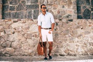 Cool and in style. Full length of handsome young African man in smart casual clothes carrying bag and looking away while standing against the stoned wall outdoors photo