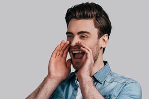 Everybody should hear it. Good looking young man in blue jeans shirt screaming and keeping hands over mouse while standing against grey background photo