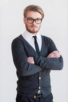 Confident young man. Handsome young man in glasses keeping arms crossed and while standing against grey background photo