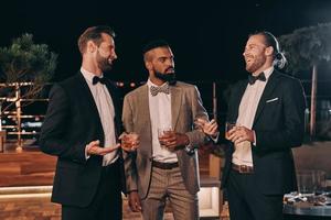 tres hombres guapos con traje bebiendo whisky y comunicándose mientras pasan tiempo en la fiesta foto