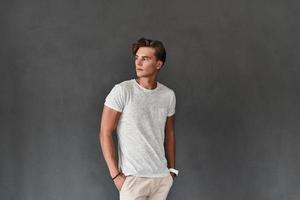 He got great style. Handsome young man looking away and keeping hands in pockets while standing against grey background photo