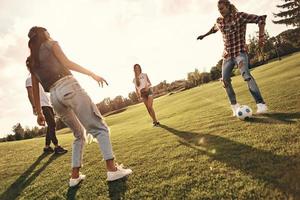 increíble juego de pies. toda la longitud de jóvenes sonrientes con ropa informal jugando al fútbol mientras están de pie al aire libre foto