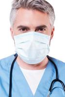 Confident surgeon. Mature grey hair doctor in surgical mask looking at camera while standing isolated on white photo