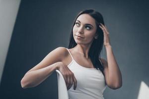Thoughtful beauty. Attractive young woman sitting on chair and adjusting her hair while sitting against grey wall photo