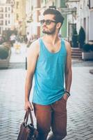 Just walking around. Handsome young man in casual wear carrying bag and looking away while walking along the street photo