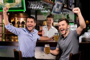 viendo la televisión en el bar. dos jóvenes felices bebiendo cerveza y gesticulando mientras están sentados en el bar foto