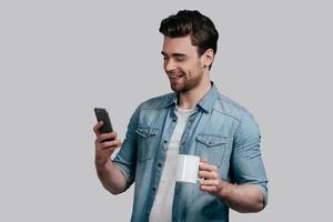 Reading interesting news. Good looking young man in blue jeans shirt holding a cup and looking at smart phone while standing against grey background photo