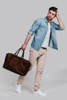 Complementing his style with bag. Full length of handsome young man in casual wear carrying brown leather bag and looking away while standing against grey background photo